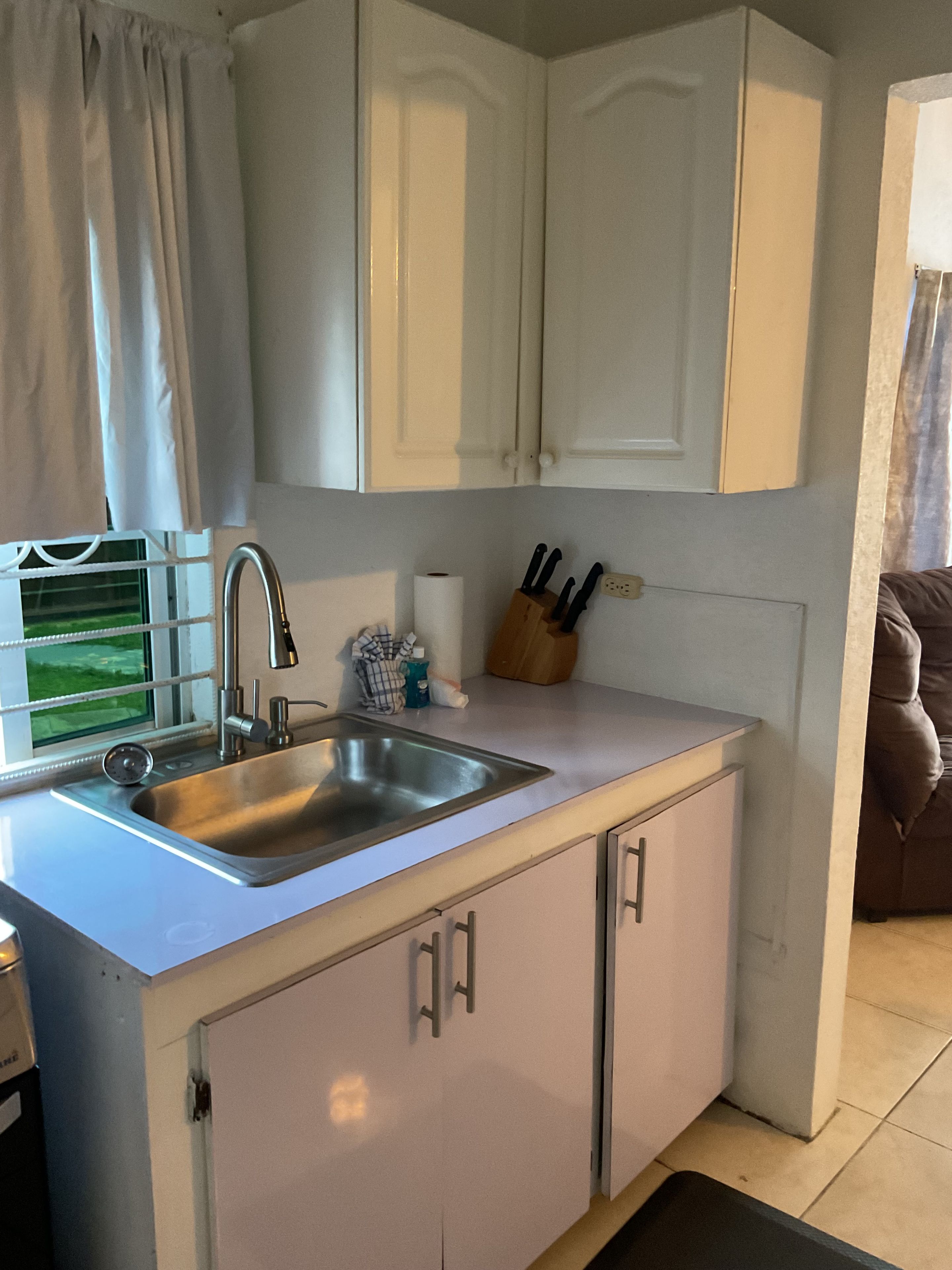 A kitchen with a stainless steel sink, white cabinets, and a window with curtains.