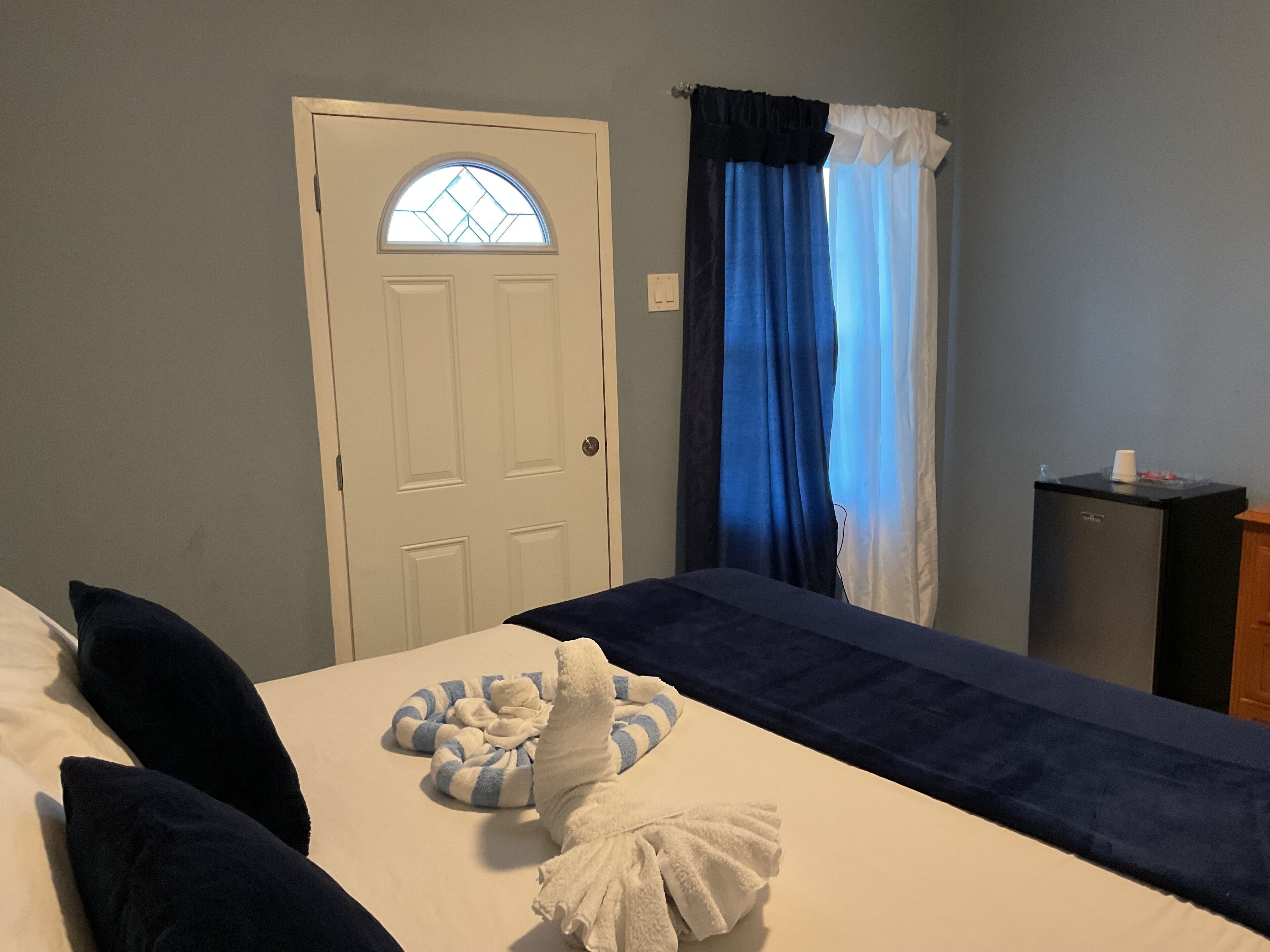 A hotel room with a bed, a small desk, a window with curtains, and a door with a glass panel.