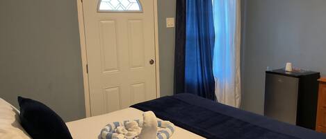A hotel room with a bed, a small desk, a window with curtains, and a door with a glass panel.