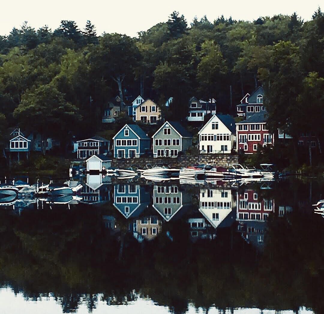 A waterfront village with houses reflected in calm water, surrounded by dense forest.