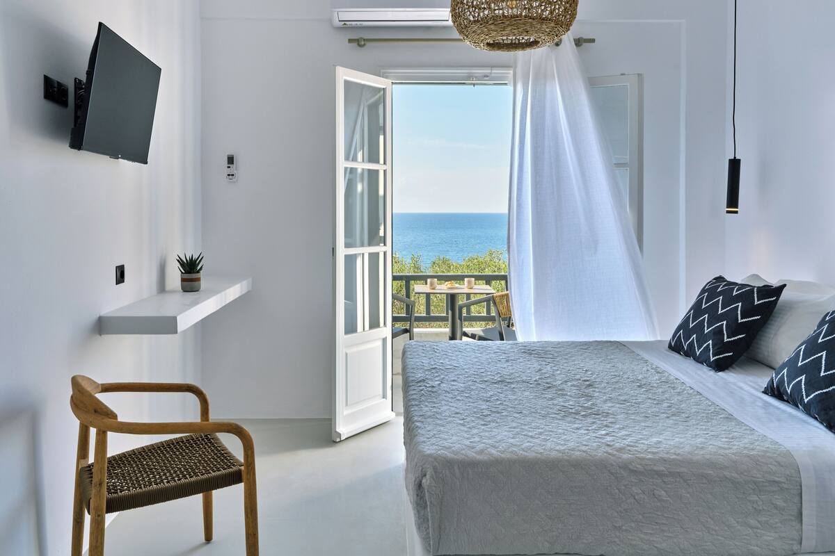 A bedroom with a bed, a chair, a TV, and a view of the sea.