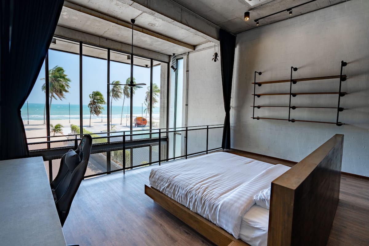 A modern bedroom with a large window overlooking a beach, a bed, a desk, and a chair.