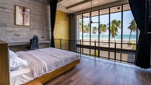 A bedroom with a large window overlooking a beach, a bed, and a concrete wall with a textured section. - The Bloc At Sea (Thap Sakae)