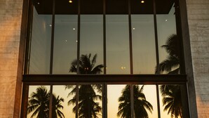 A modern building with large windows reflecting palm trees and a sunset over the ocean. - The Bloc At Sea (Thap Sakae)