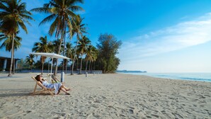 A beach with a person relaxing on a lounge chair under a parasol, palm trees, and a clear sky. - The Bloc At Sea (Thap Sakae)