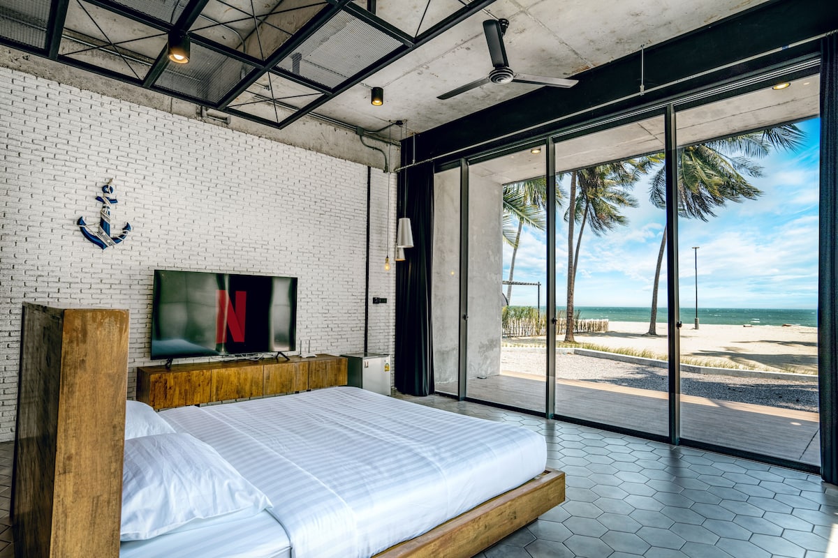 A modern bedroom with a large bed, a TV, and a view of the beach.