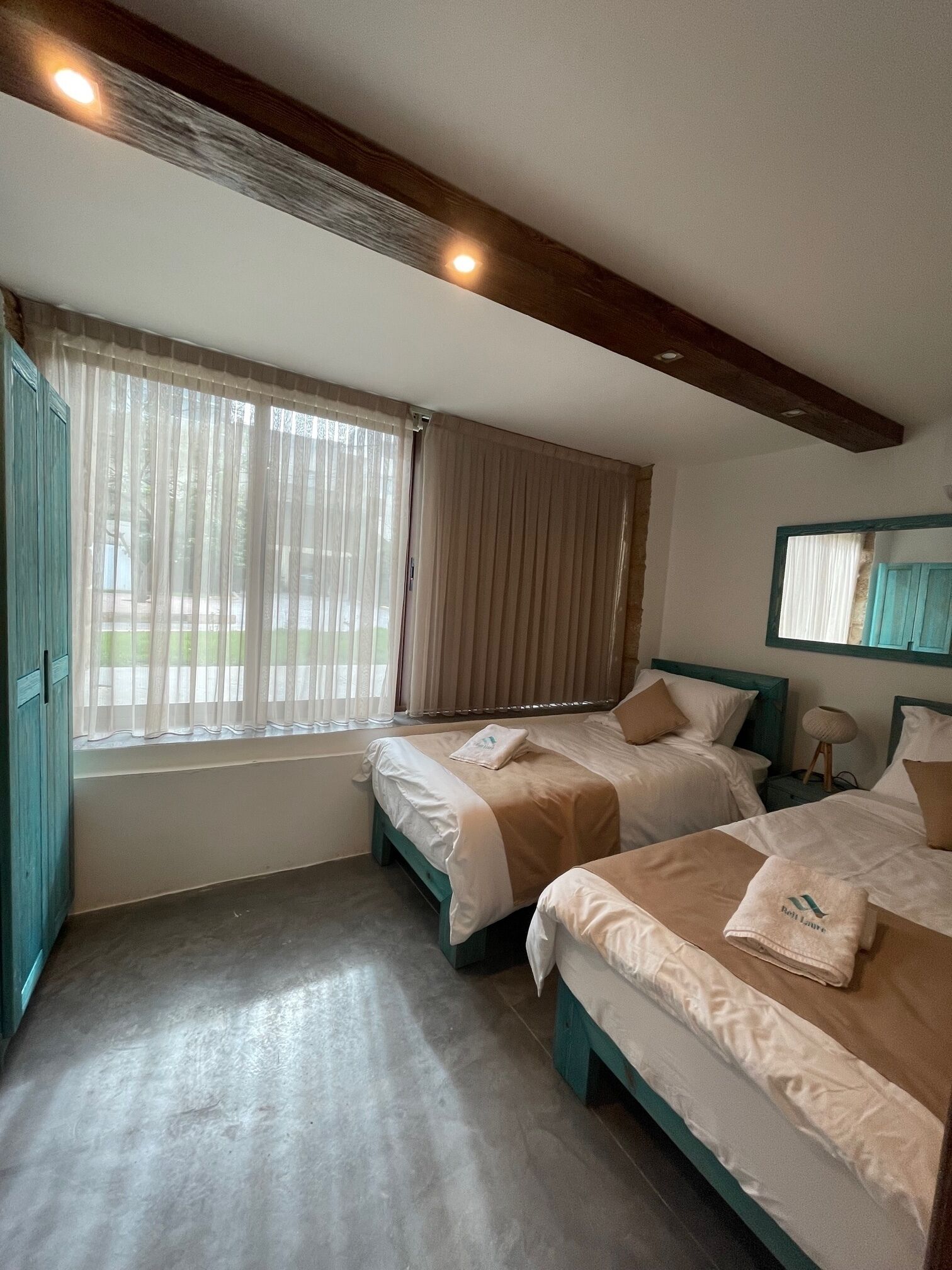Un dormitorio con dos camas, una ventana con cortinas, techo de madera y una mesita de noche con lámpara.