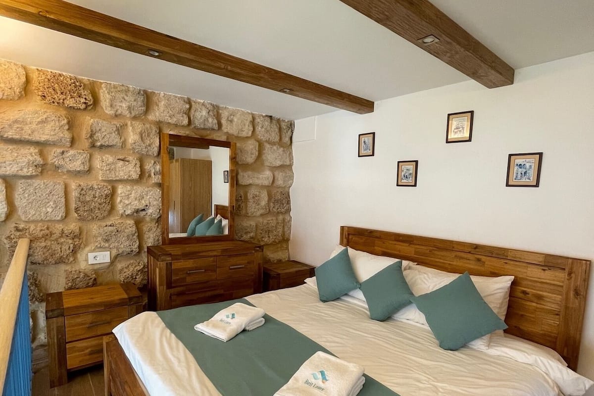 Un dormitorio con pared de piedra, vigas de madera, una cama grande con ropa de cama verde y blanca, y una mesita de noche de madera.