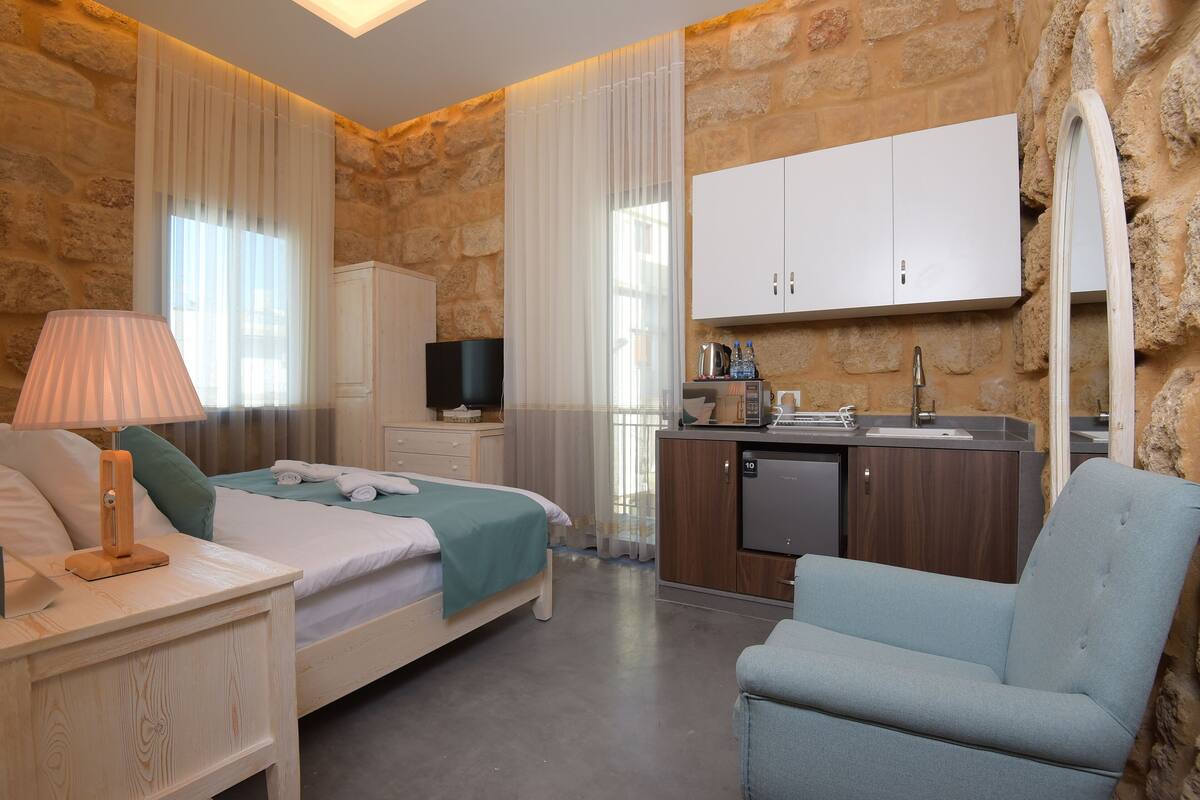 Un dormitorio con cama, mesita de noche, lámpara y una silla. Hay una kitchenette con lavabo, estufa y armarios.