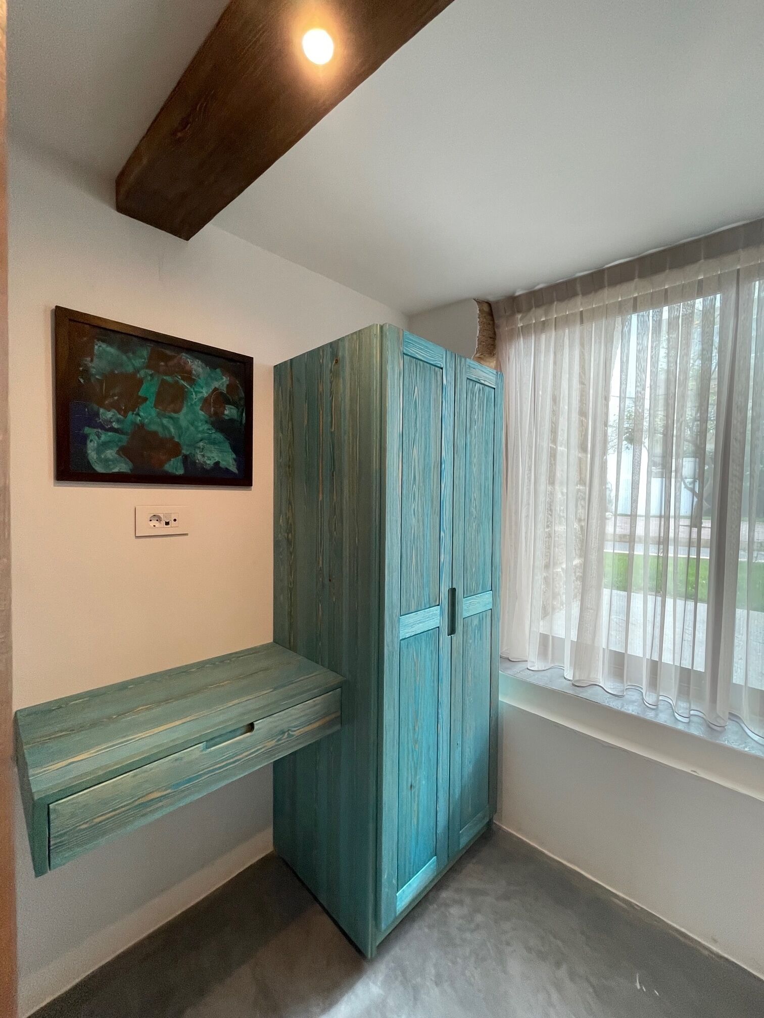 Una habitación con un armario verde azulado, un banco, un cuadro y una ventana con cortinas de gasa.