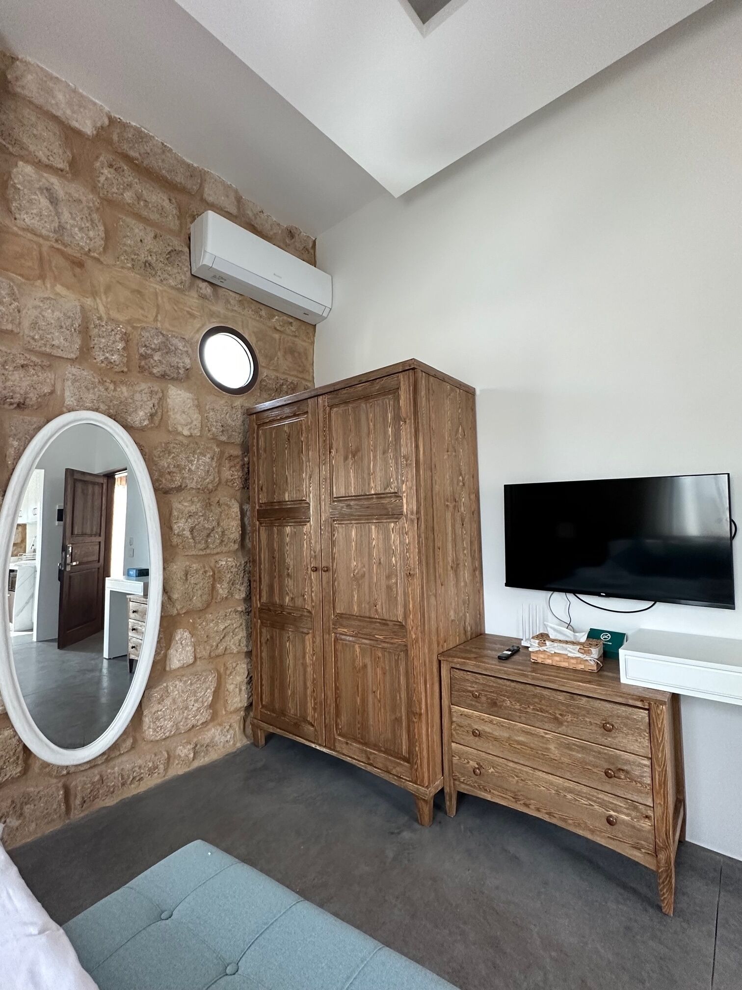 Una habitación con pared de piedra, un armario de madera, un espejo, un televisor y una cómoda de madera pequeña.