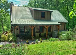 Una casa in legno a due piani con tetto in metallo, un portico con travi in legno e un giardino con diverse piante e fiori.