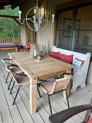 Een houten terras met een grote tafel en rieten stoelen, een bank met rode kussens en een kroonluchter.