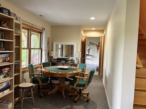 Een gezellige kamer met een ronde houten tafel, omringd door groene fauteuils, een boekenplank vol boeken en een grote spiegel erboven.