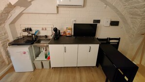 Eine kleine Küche mit einem weißen Kühlschrank, einem Herd, einer Spüle und einer schwarzen Arbeitsplatte. Es befindet sich ein weißer Schrank mit zwei Türen und ein schwarzer Stuhl dort. Eine Klimaanlage ist über der Arbeitsplatte an der Wand angebracht.