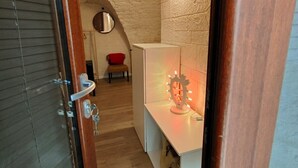 Ein schmaler Flur mit einer Holztür, einer weißen Wand mit strukturierter Oberfläche, einem kleinen roten Stuhl und einem weißen Schrank mit einer Leuchte.