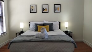 Un lit bien fait, avec une serviette pliée, une table de chevet avec une lampe et trois œuvres d’art encadrées accrochées au mur.