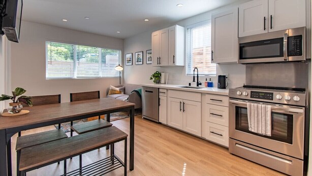 A modern kitchen with white cabinets, stainless steel appliances, a dining table with benches, and a TV mounted on the wall. - Modern Seattle Townhome Near Downtown & Parks (Seattle)