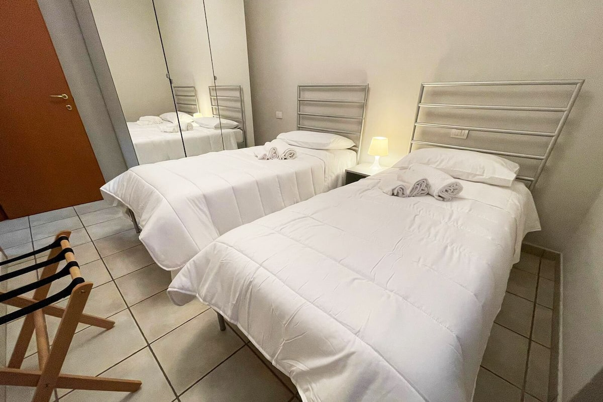 Ein Hotelzimmer mit zwei Betten, jeweils mit Kopfteil, einem Holzstuhl und einem Nachttisch mit Lampe.