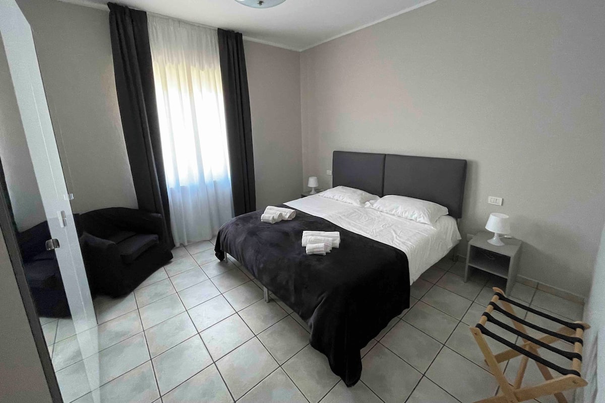 Ein modernes Hotelzimmer mit einem großen Bett, einem Nachttisch mit Lampe, einem Sessel und einem hölzernen Wäscheständer.