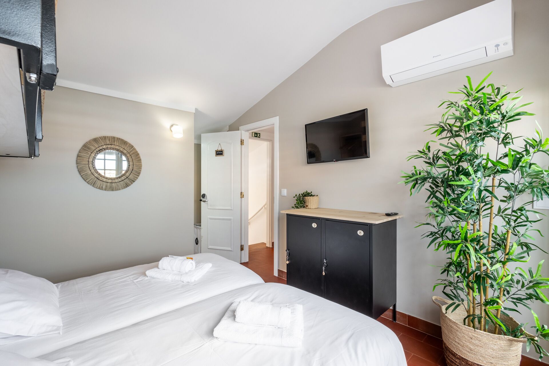 A hotel room with two beds, a TV, a wall-mounted air conditioner, a mirror, and a potted plant.