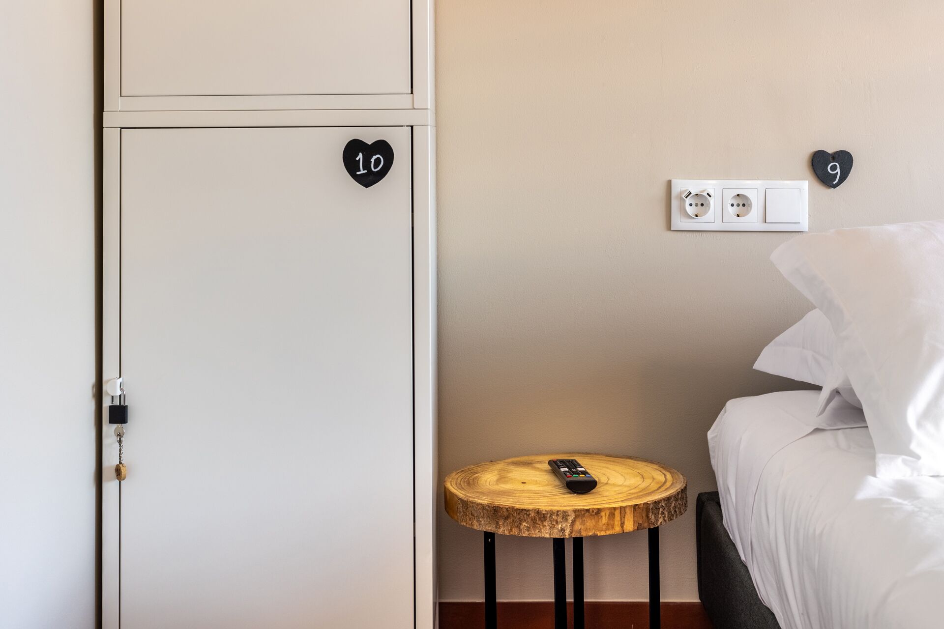 Un dormitorio con una cama, una mesita de noche de madera con un control remoto, un enchufe y una puerta con un letrero con forma de corazón que indica el número 10.