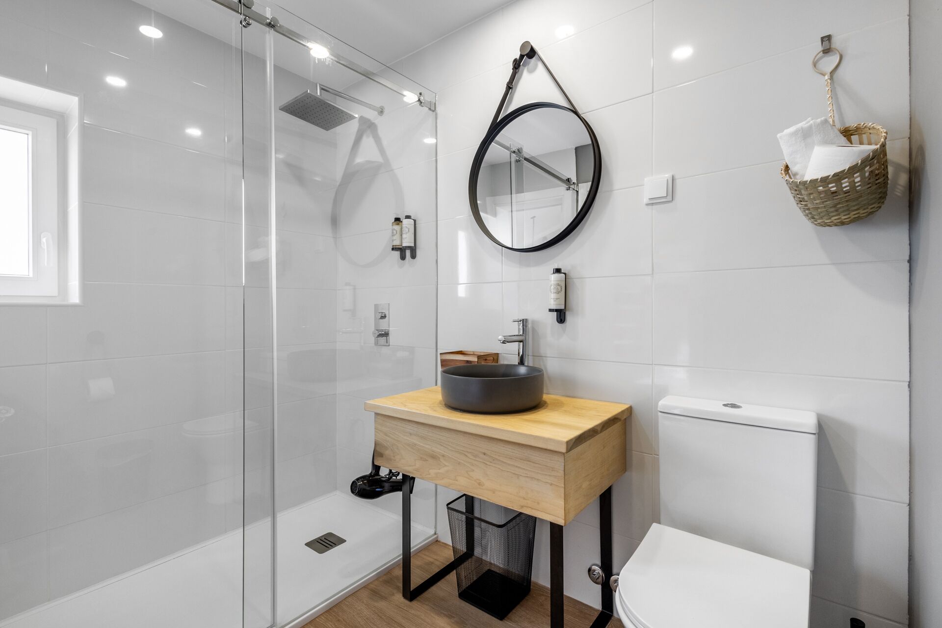 A modern bathroom with a glass-enclosed shower, a round mirror, a wooden vanity with a vessel sink, and a toilet.