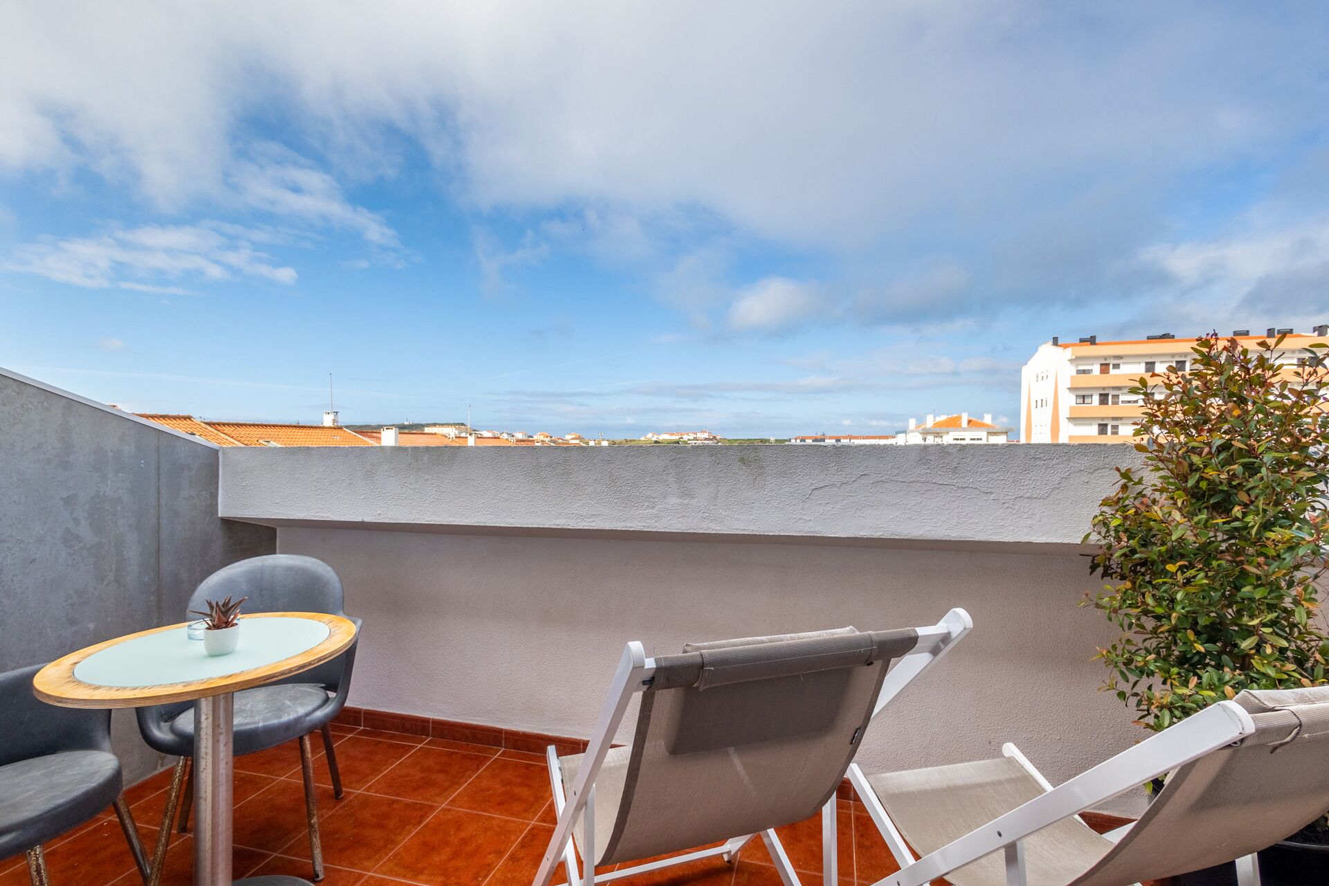 Terraza en la azotea con una mesa, dos sillas, una planta en maceta y vistas a edificios distantes.