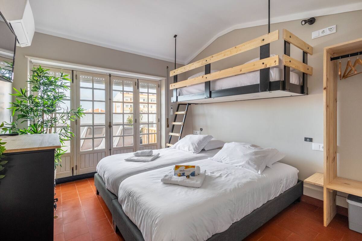 A bedroom with a bed, a wooden loft bed, a plant, a window, and a door.