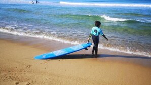 Une plage de sable où un enfant, tenant une planche de surf, marche en direction de l’océan.