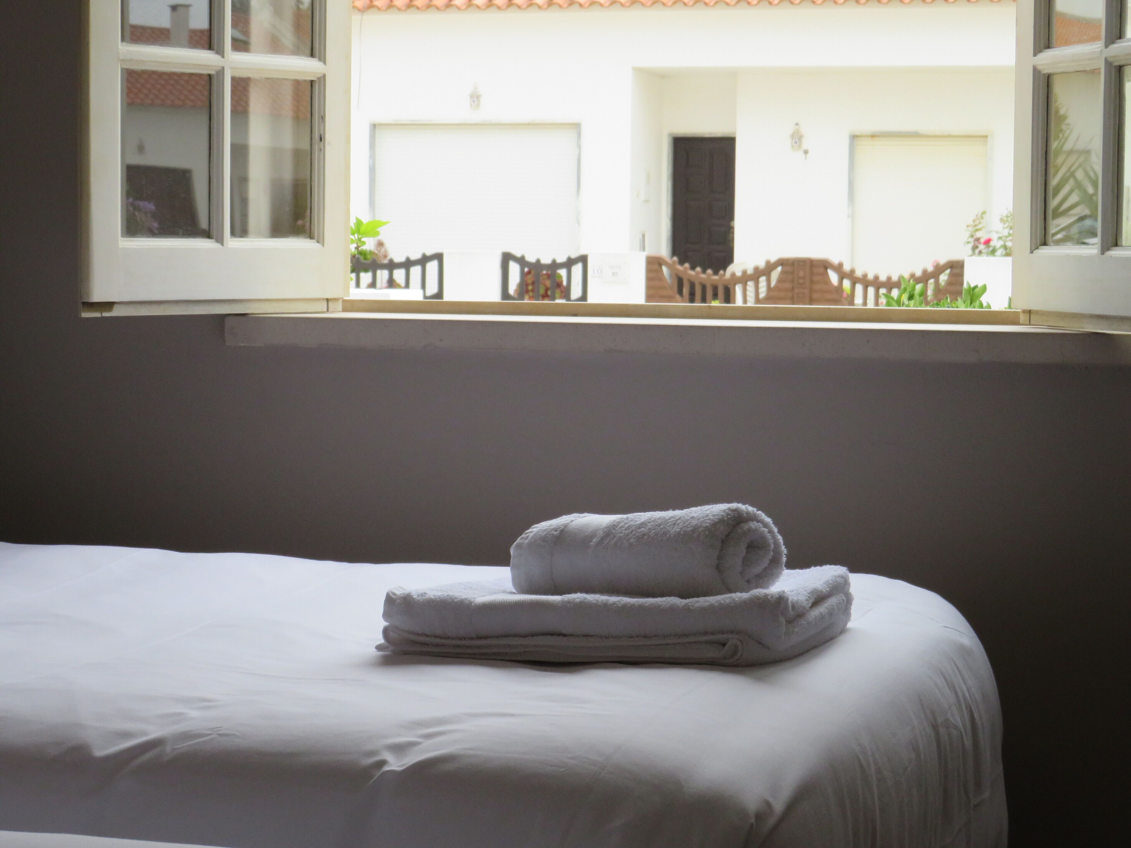 A neatly made bed with a folded towel, a window with a view of an outdoor patio area with chairs and plants.