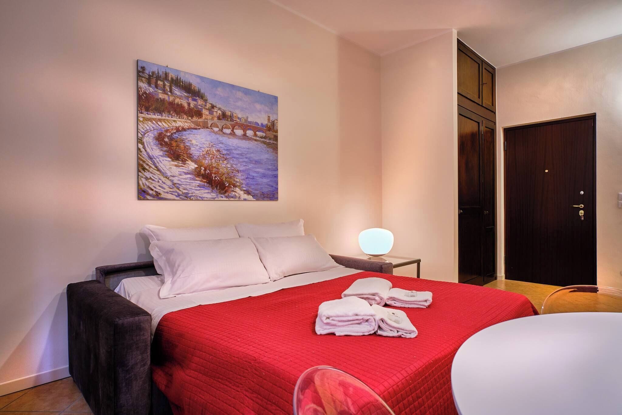 A hotel room with a bed, a painting on the wall, a bedside table with a lamp, and a door.