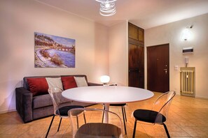 A modern living room with a round dining table, clear chairs, a grey sofa with red and white cushions, a painting on the wall, and a wooden door. - Suite Mery (Verona)