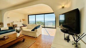 A modern living room with a large TV, comfortable seating, and a view of the landscape through large windows. - Cardina CostaBravaSi - Pool, A/C, barbecue, views (Sant Feliu de Guíxols)