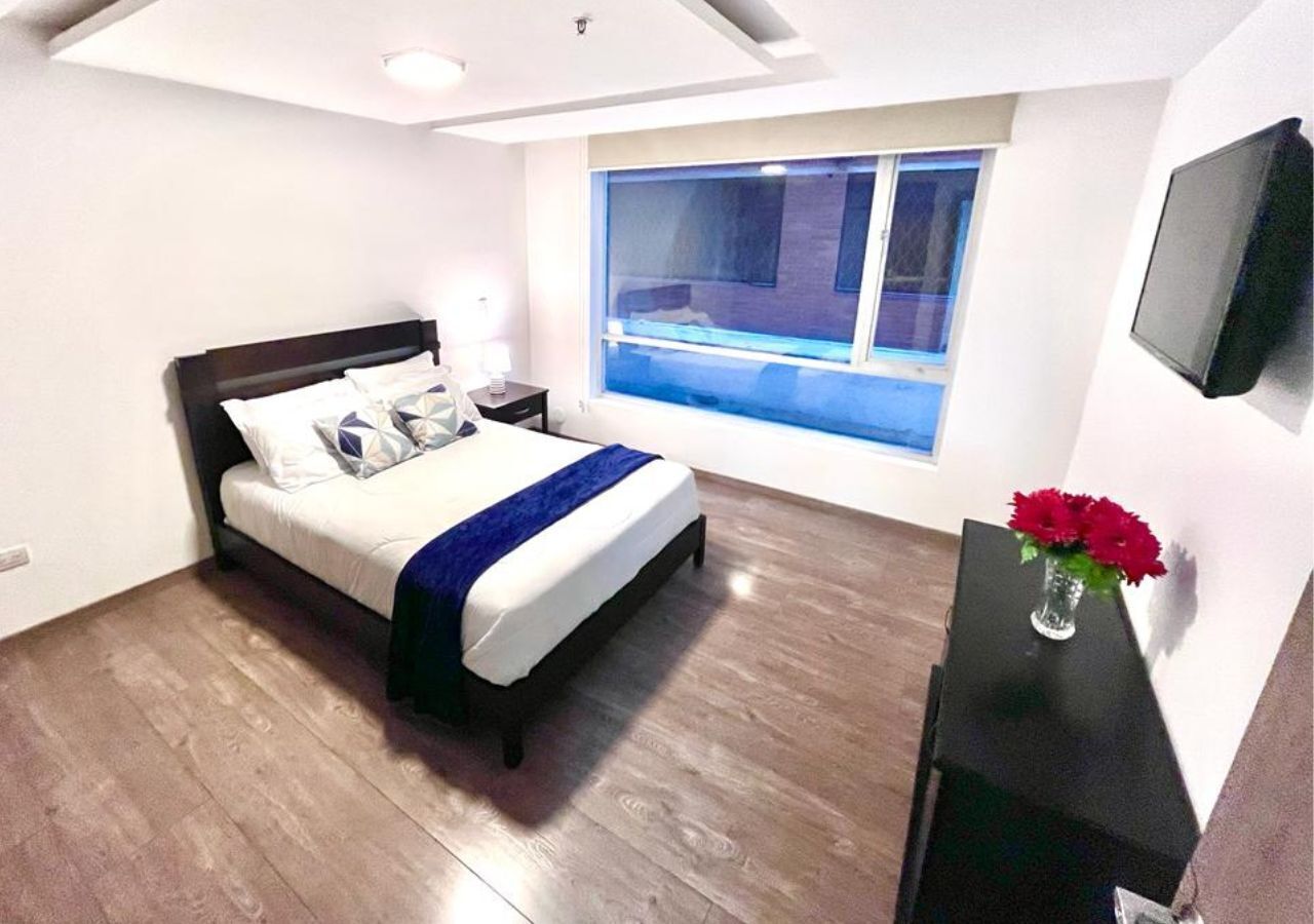 Quarto moderno com uma cama grande, mesa de cabeceira, escrivaninha e janela com vista para o exterior.