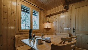 A wooden interior with a dining table, chairs, a window with a view, and a shelf with a lamp.