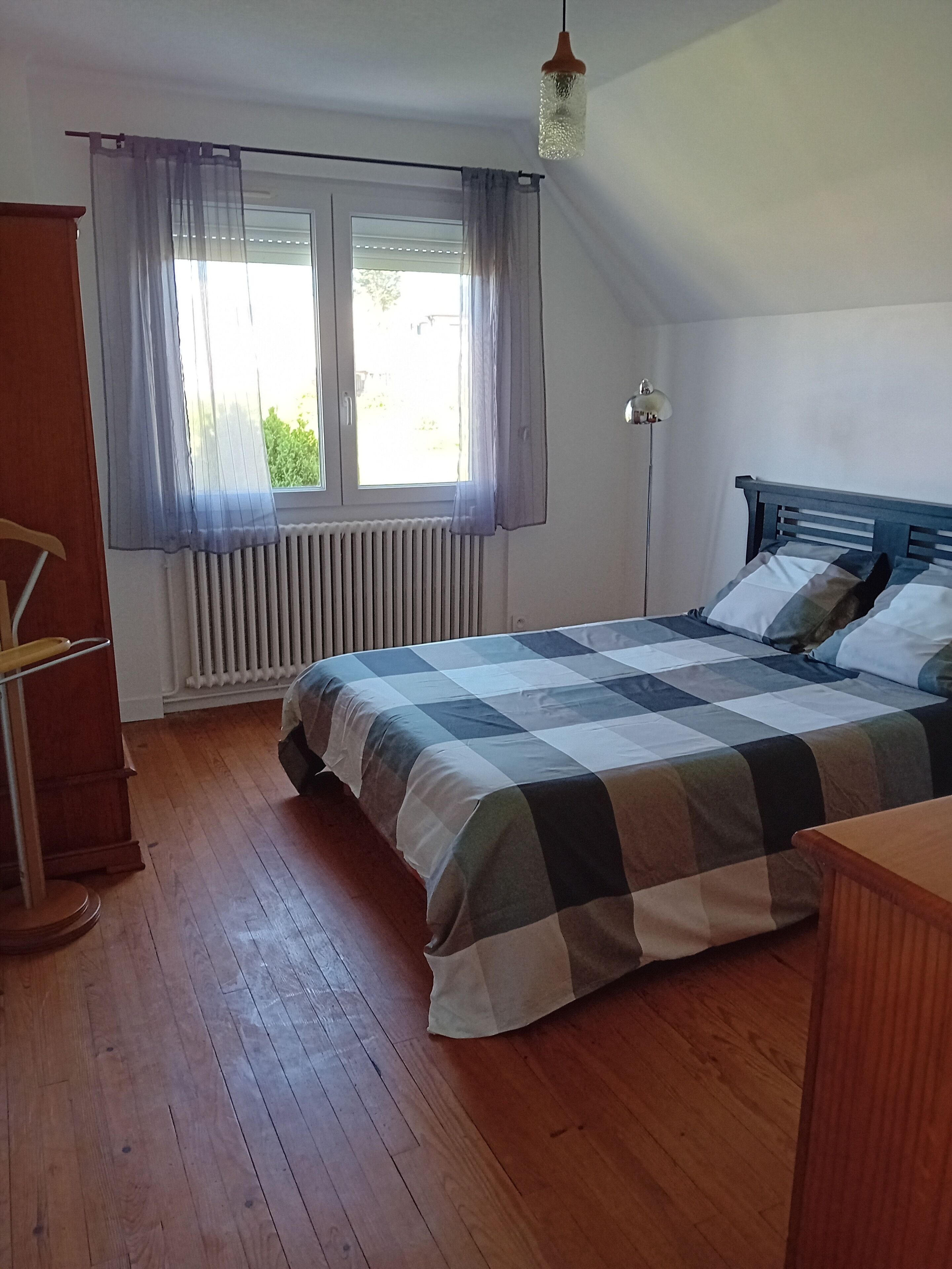 A bedroom with a checkered bedspread, a wooden floor, a radiator, a window with sheer curtains, and a hanging light fixture.
