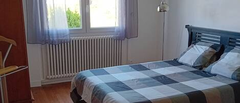 A bedroom with a checkered bedspread, a wooden floor, a radiator, a window with sheer curtains, and a hanging light fixture.