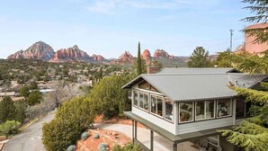 Una casa moderna con un amplio balcón de vidrio, rodeada de vegetación desértica y montañas al fondo.