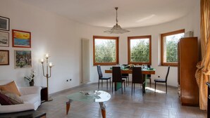 Amplia sala de estar con una mesa de centro de vidrio, un comedor con mesa y sillas de madera, y ventanas grandes con vistas a zonas verdes.