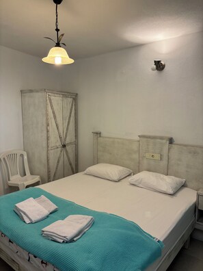 A single bed with white bedding, a teal blanket, and two folded towels. A white plastic chair is to the left.