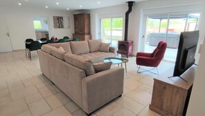 Amplia sala de estar con un sofá color beige, un sillón rojo, una mesa de centro de vidrio y un televisor. Hay una zona de comedor con sillas verdes y un armario de madera.