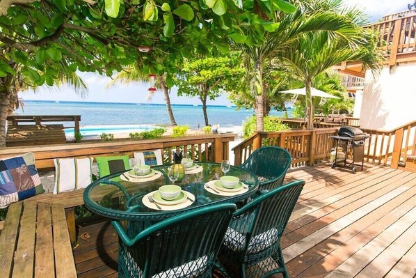Une terrasse en bois avec une table en verre dressée pour le repas, entourée de chaises vertes, donnant sur une plage bordée de palmiers et offrant une vue dégagée sur l’océan.