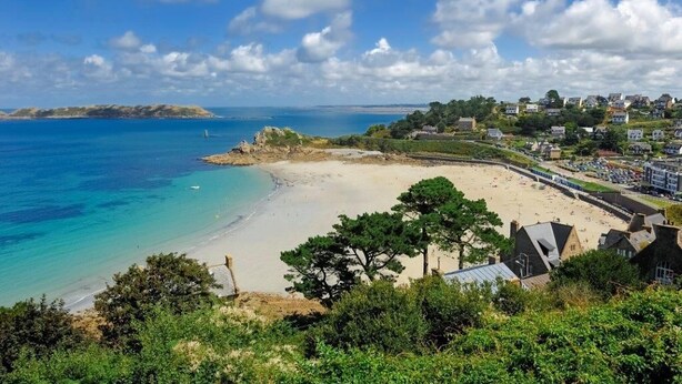 A coastal town with a sandy beach, a clear blue sea, and a hilly backdrop with residential buildings.