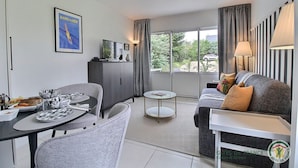 A modern living room with a dining area, a grey sofa, a small table, and a cabinet. There is a large window with curtains, a framed picture on the wall, and a logo on the wall.