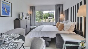 A hotel room with a bed, a desk, a chair, a small table, a lamp, a TV, and a poster on the wall.