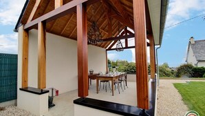 A covered outdoor dining area with a table and chairs, wooden beams, and a view of the surrounding landscape.