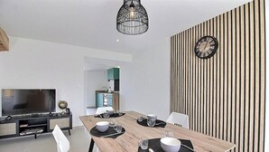 Un comedor moderno con una mesa de madera dispuesta para cuatro, un reloj blanco y negro en la pared y un televisor al fondo.