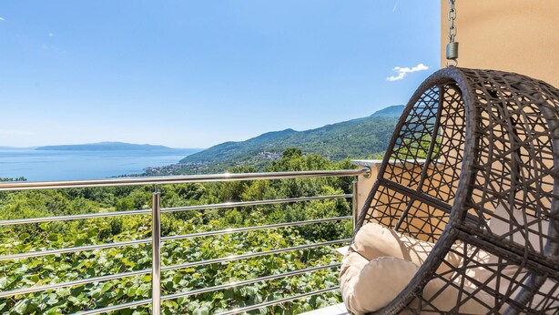 Un balcone con ringhiera metallica, una sedia in vimini e vista su un paesaggio costiero con montagne e un corpo d'acqua.