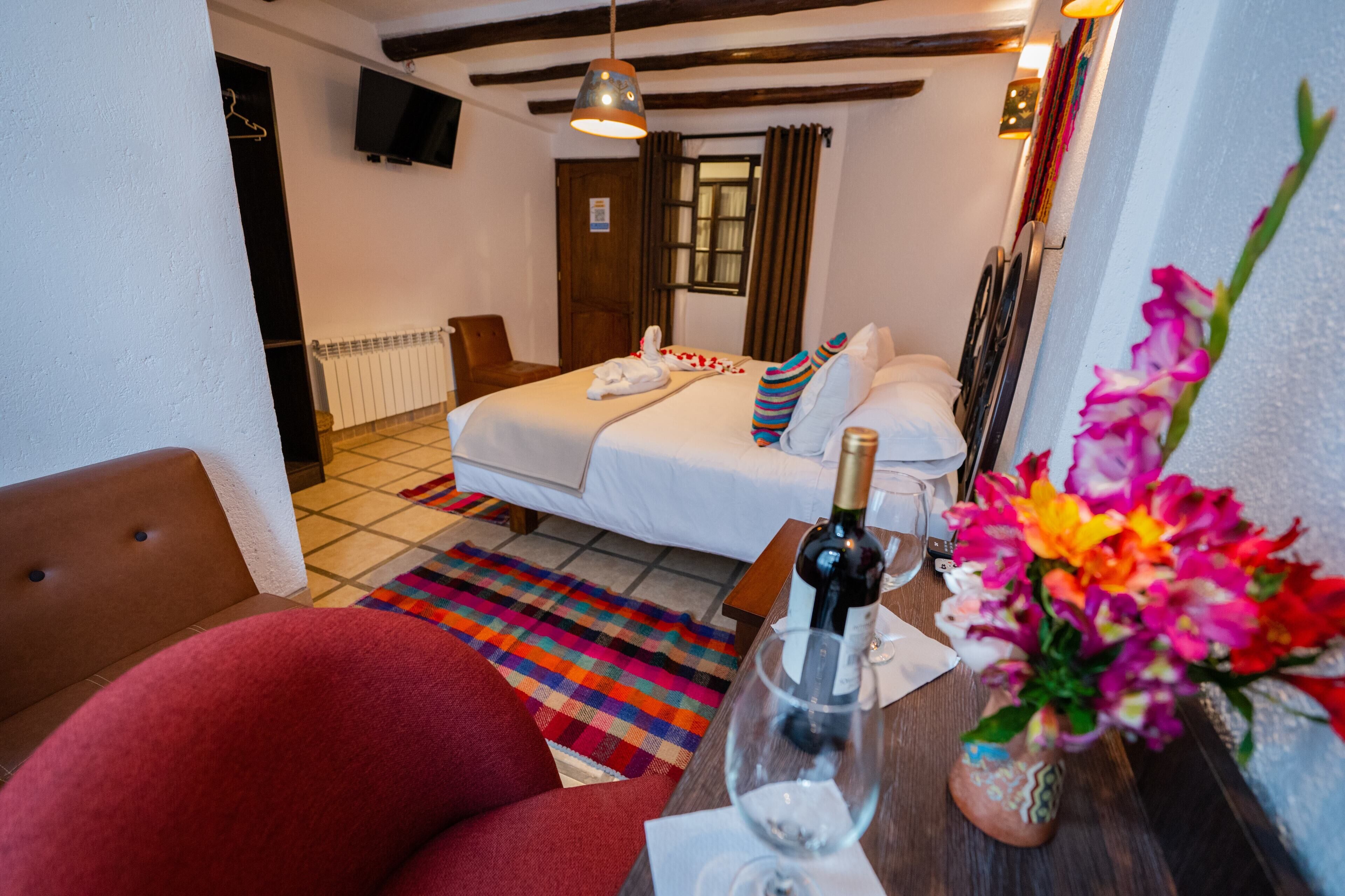 Habitación de hotel con una cama, un sillón rojo, una mesa de madera con una botella y un vaso, y una alfombra colorida.